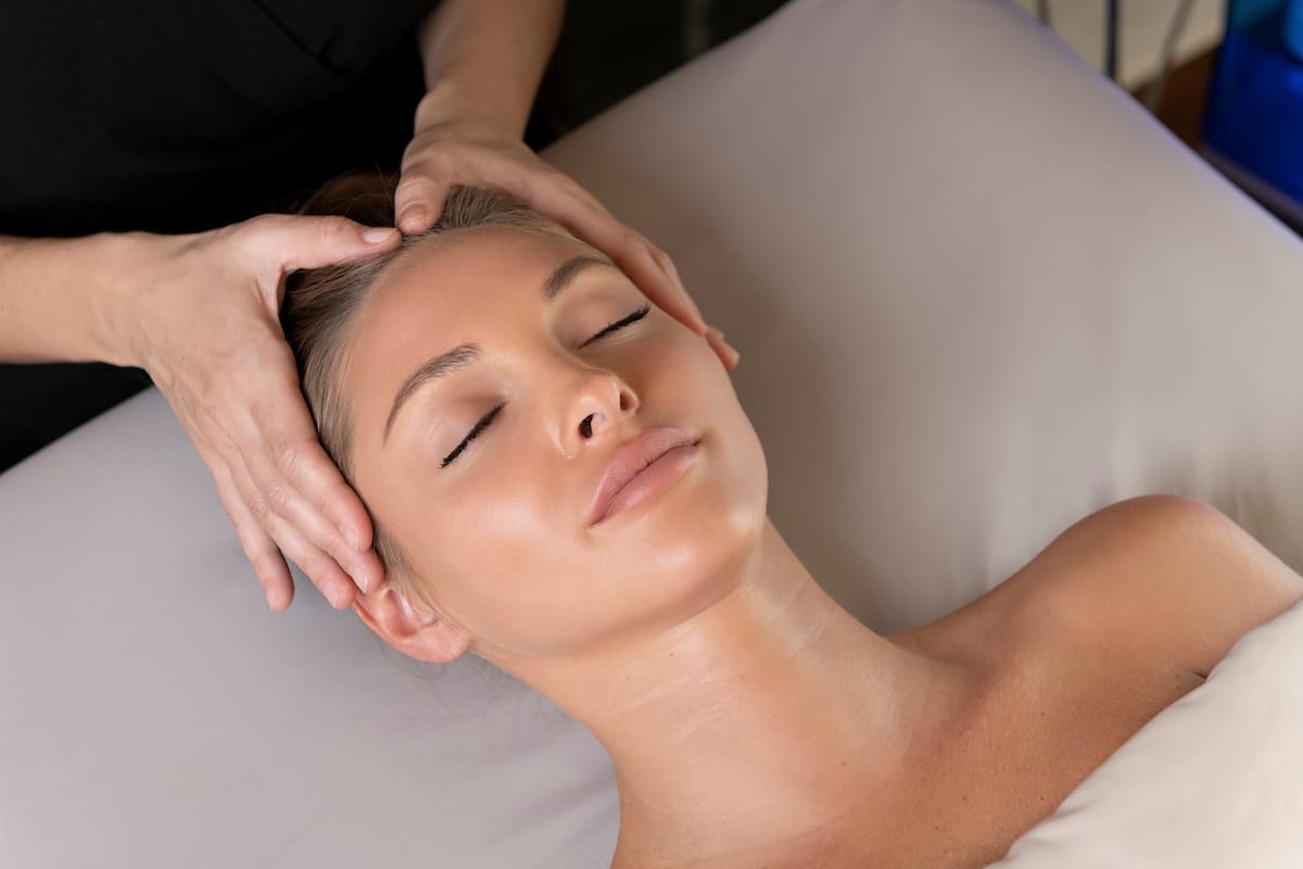 Close view of a woman's face, eyes closed, on a cream cushion while a therapist's hands press her temples and scalp.